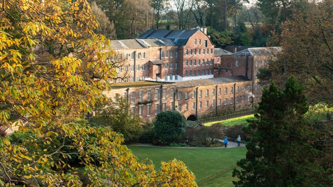An elevated view of a red brick mill building, peaking out amongst trees in autumn leaf